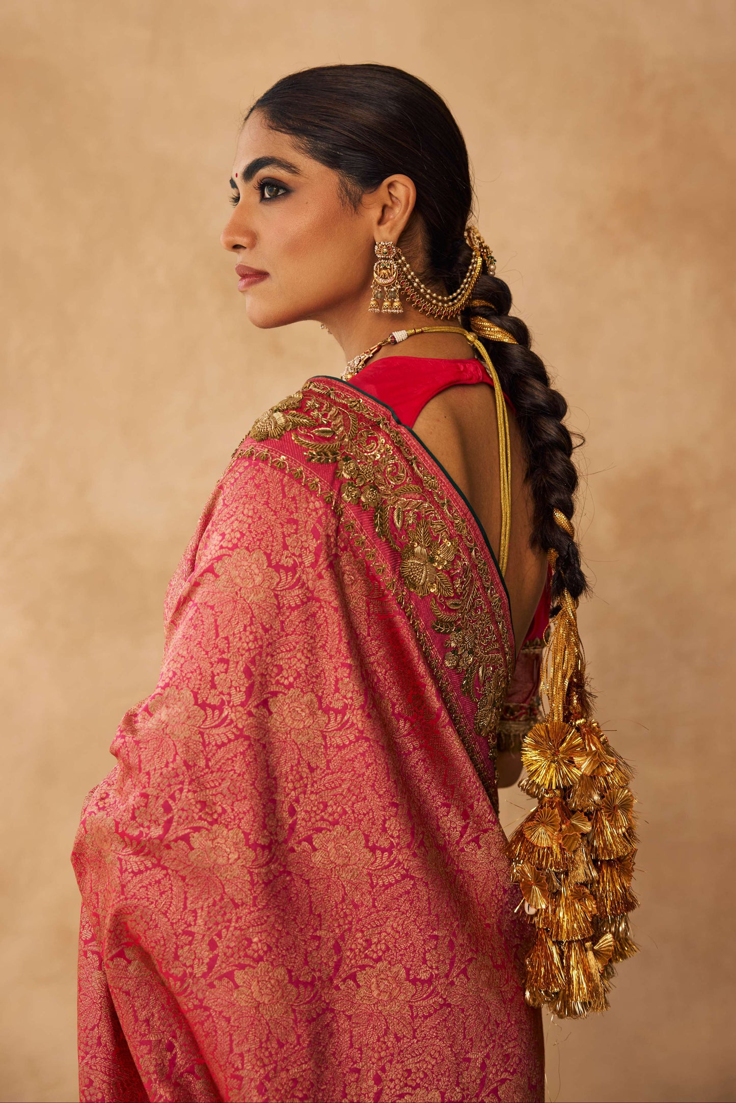 Woman in a pink saree with gold jewelry against a beige background